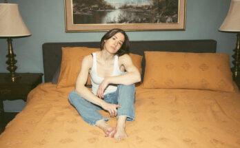 Promotional photo of Laura Stevenson sitting on an orange bed while wearing a white t-shirt and blue jeans.