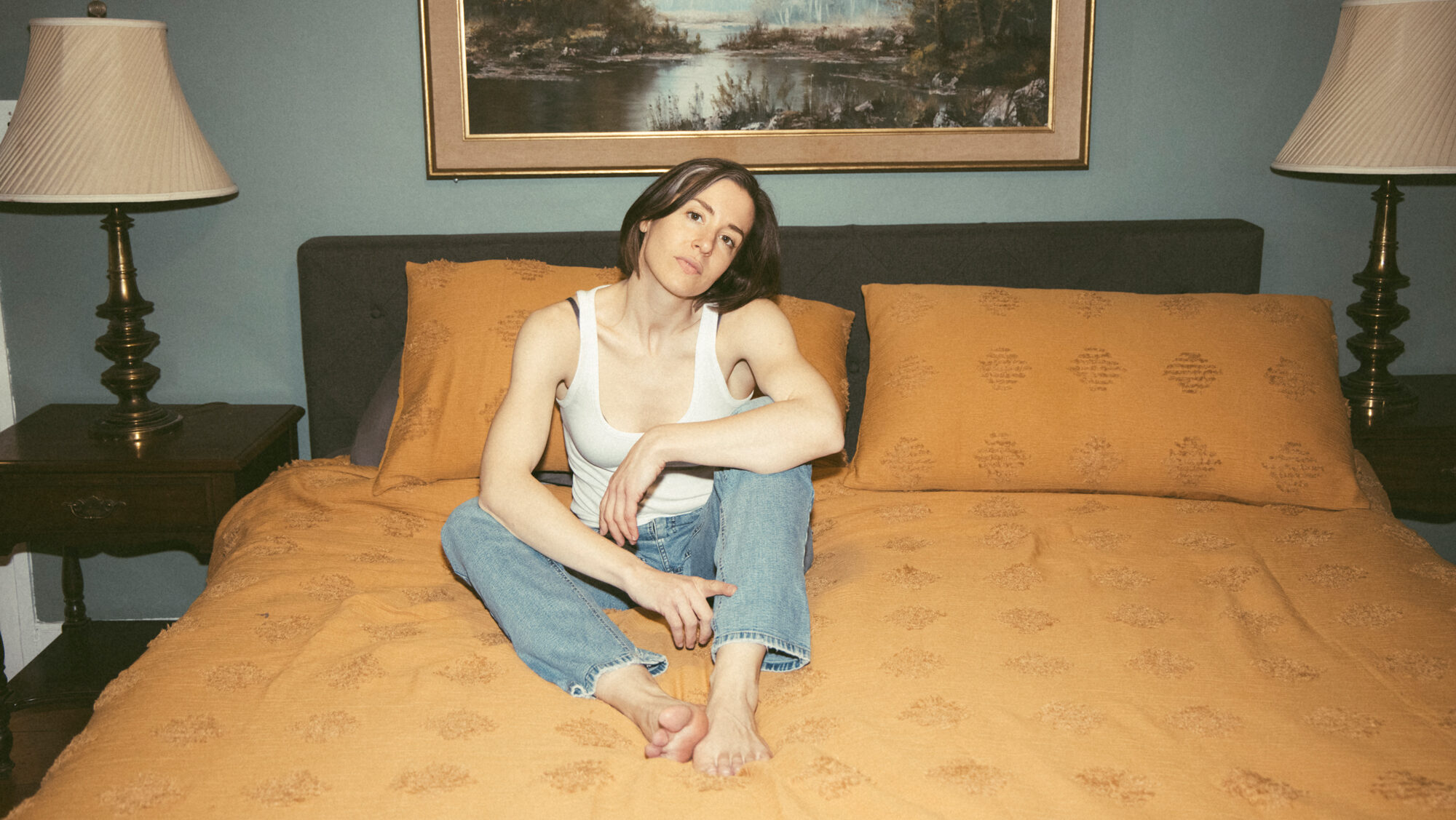 Promotional photo of Laura Stevenson sitting on an orange bed while wearing a white t-shirt and blue jeans.